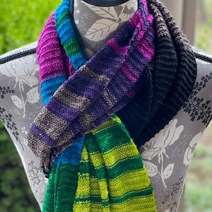 May include: A colorful striped knitted scarf with shades of pink, purple, blue, green, and black. The scarf is folded and draped over a mannequin.