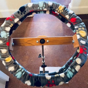 Może przedstawiać: Sztywny obręcz z tkaniny w kolorze szarym i białym z kolorowym wzorem, zamontowany na drewnianym stojaku z regulowanym metalowym ramieniem. Obręcz jest przeznaczony do haftu lub innych prac ręcznych.