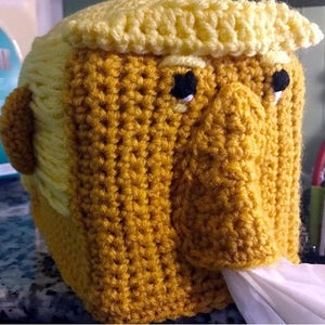 May include: A crocheted tissue box cover shaped like a person with yellow hair and a brown face. The tissue box cover has a small opening for the tissues to come out.