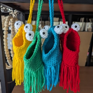 May include: Four brightly coloured crochet monster pouches hang from a metal rack. Each pouch is a different colour: yellow, green, blue, and red. They feature large, white, googly eyes and fringe at the bottom. The pouches are suspended by a matching crochet cord.