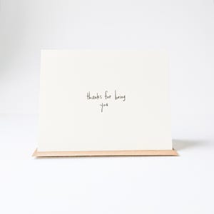 Può includere: Un biglietto di auguri quadrato, bianco sporco, con la scritta a mano "thanks for being you" in inchiostro nero. Il biglietto è appoggiato su una piccola base rettangolare marrone, su sfondo bianco.