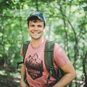 May include: A man wearing a pink t-shirt with a mountain and trees graphic and the text "Explore More" is smiling at the camera. He is wearing a green backpack and a dark green baseball cap. The background is a blurred image of green trees.