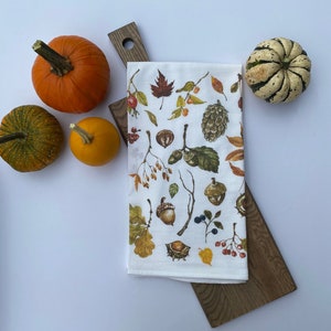 May include: A white kitchen towel with a fall-themed print of leaves, acorns, berries, and pine cones. The towel is folded and placed on a wooden cutting board. There are three small pumpkins and one larger pumpkin in the background.