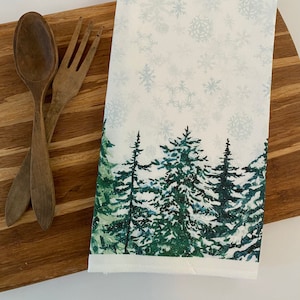 May include: White kitchen towel with a snowy forest scene. The towel features a green pine forest with a white snowy background and a light blue snowflake pattern.
