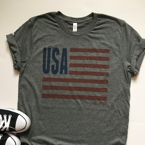 Pode incluir: Uma camiseta cinza com um gr&aacute;fico de bandeira americana azul e vermelha e o texto "USA est. 1776".