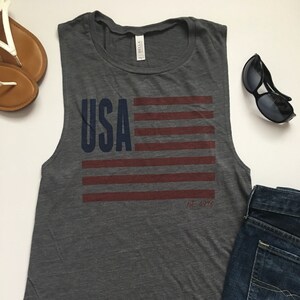 K&ouml;nnte beinhalten: Graues &auml;rmelloses Tanktop mit einem rot-wei&szlig;-blauen amerikanischen Flaggen-Motiv und dem Text "USA est. 1776".
