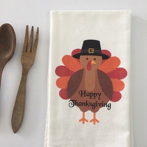 Puede incluir: Una toalla de cocina blanca con un dibujo de un pavo que lleva un sombrero de peregrino. El pavo es marr&oacute;n con plumas naranjas y rojas. El texto "Happy Thanksgiving" est&aacute; impreso debajo del pavo.