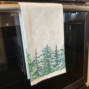 May include: A white kitchen towel with a winter forest scene. The towel features a watercolor-style design of green pine trees with snowflakes on a light blue background.