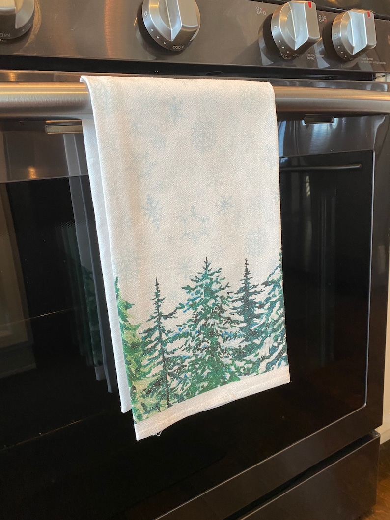 May include: A white kitchen towel with a winter forest scene. The towel features a watercolor-style design of green pine trees with snowflakes on a light blue background.