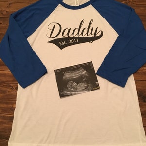 Puede incluir: Camiseta de estilo b&eacute;isbol blanca y azul con el texto "Daddy Est. 2017" en negro. Una imagen de ultrasonido en blanco y negro est&aacute; en la camiseta.