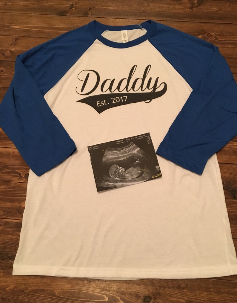Puede incluir: Camiseta de estilo b&eacute;isbol blanca y azul con el texto "Daddy Est. 2017" en negro. Una imagen de ultrasonido en blanco y negro est&aacute; en la camiseta.