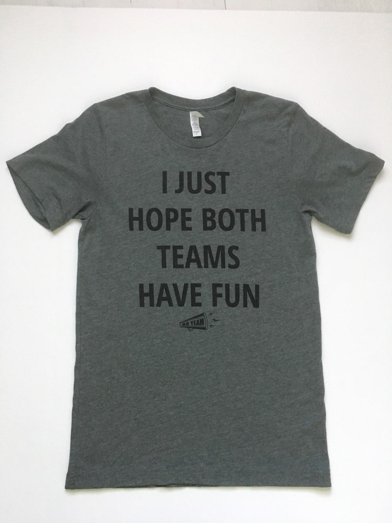 May include: A heather gray t-shirt with the text "I just hope both teams have fun" printed in black. A small black and white illustration of a megaphone with the text "Go Team" is below the text.