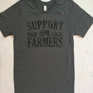 May include: A dark grey t-shirt with the text "Support Your Local Farmers" printed in black. A small tractor graphic is included in the text.