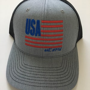 May include: A grey and black trucker hat with a red, white and blue American flag design and the text "USA est. 1776".