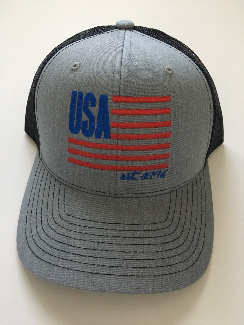 May include: A grey and black trucker hat with a red, white and blue American flag design and the text "USA est. 1776".