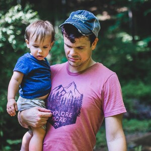 May include: A man wearing a pink t-shirt with a mountain and trees graphic and the text "Explore More" holds a young child in his arms. The man is wearing a blue baseball cap.