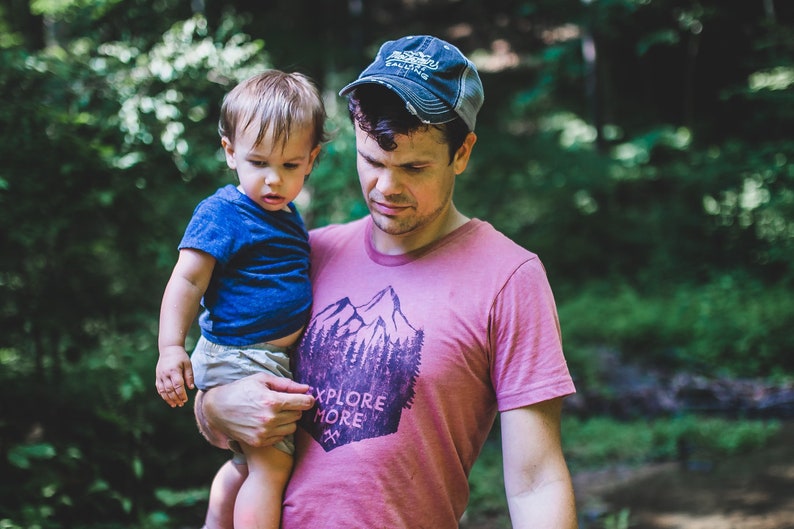 May include: A man wearing a pink t-shirt with a mountain and trees graphic and the text "Explore More" holds a young child in his arms. The man is wearing a blue baseball cap.