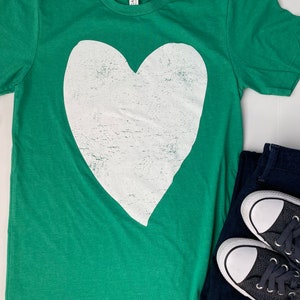 Könnte beinhalten: Ein grünes T-Shirt mit einem großen weißen Herz-Motiv. Das Herz ist abgenutzt und hat einen Vintage-Look. Das Shirt liegt flach auf einer weißen Oberfläche.