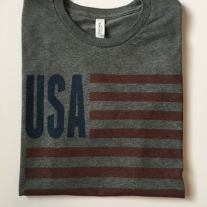 Pode incluir: Uma camiseta cinza mescla com um design de bandeira americana vermelha, branca e azul e a palavra "USA" em azul marinho.