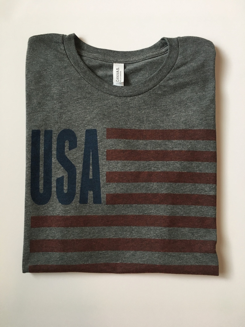 Pode incluir: Uma camiseta cinza mescla com um design de bandeira americana vermelha, branca e azul e a palavra "USA" em azul marinho.
