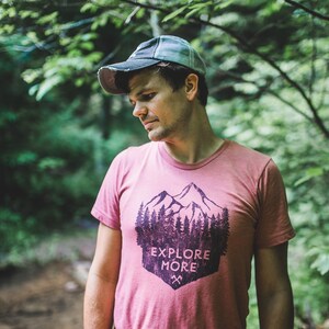 May include: A man wearing a pink t-shirt with a mountain and trees graphic and the text "Explore More". He is wearing a grey and black baseball cap and is standing in a forest.