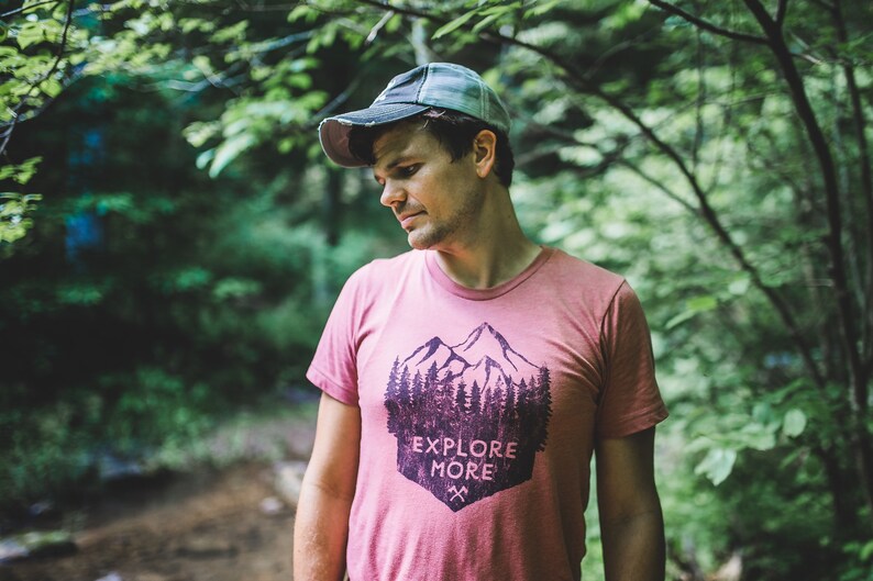 May include: A man wearing a pink t-shirt with a mountain and trees graphic and the text "Explore More". He is wearing a grey and black baseball cap and is standing in a forest.