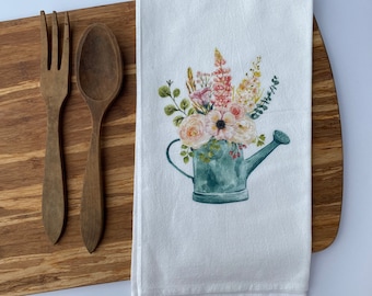 Könnte beinhalten: Ein weißes Geschirrtuch mit einer Aquarell-Illustration einer grünen Gießkanne, gefüllt mit rosa, weißen und gelben Blumen.