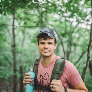 May include: A man wearing a pink t-shirt with a mountain graphic and a gray baseball cap is hiking in a forest. He is carrying a green backpack and holding a blue water bottle.