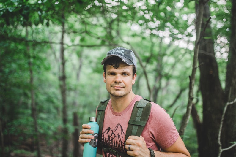 May include: A man wearing a pink t-shirt with a mountain graphic and a gray baseball cap is hiking in a forest. He is carrying a green backpack and holding a blue water bottle.
