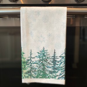 May include: A white kitchen towel with a winter forest scene printed on it. The towel features a snowy background with green pine trees and snowflakes.