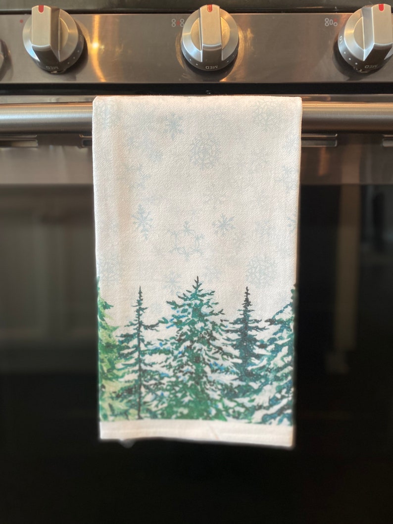 May include: A white kitchen towel with a winter forest scene printed on it. The towel features a snowy background with green pine trees and snowflakes.