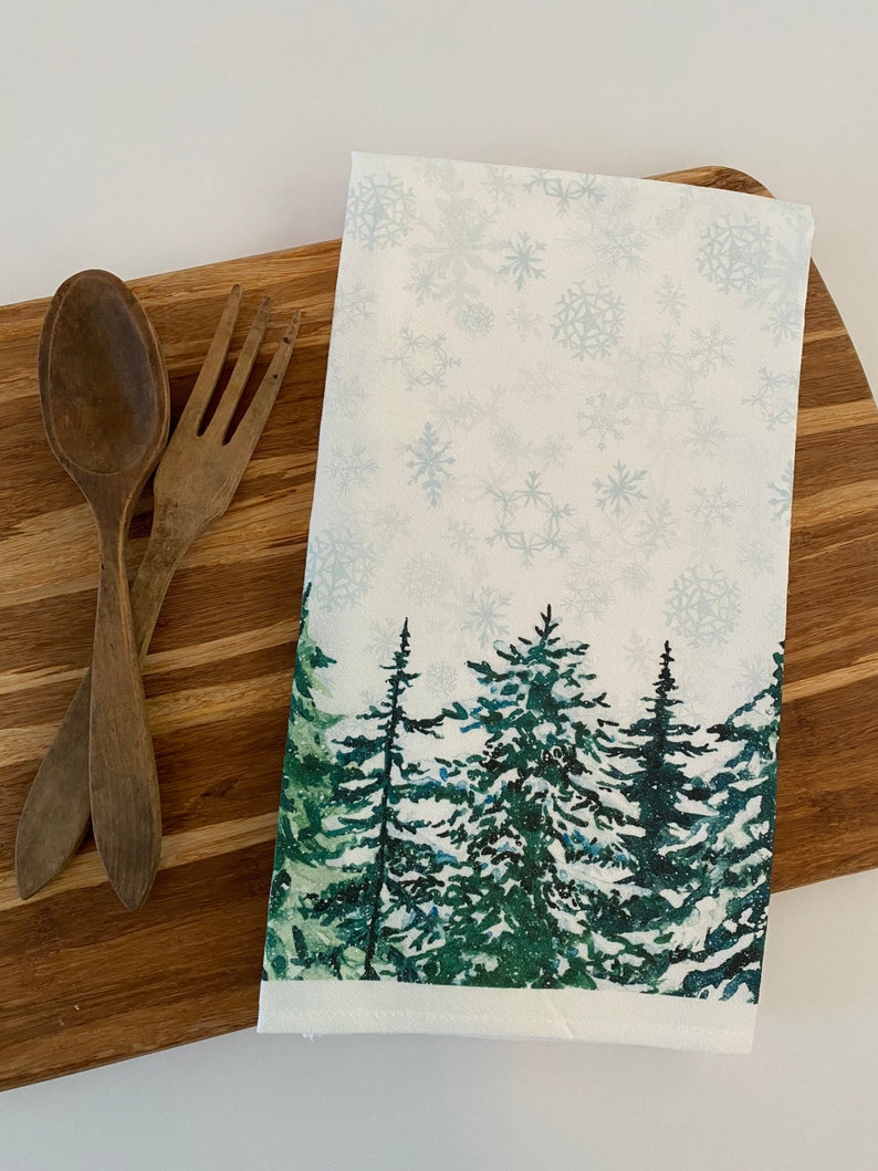 May include: White kitchen towel with a snowy forest scene. The towel features a green pine forest with a white snowy background and a light blue snowflake pattern.