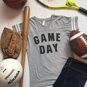 May include: A gray t-shirt with the text "GAME DAY" printed on the front. The shirt has white stripes on the sleeves. The image also includes a baseball bat, a baseball glove, a basketball, a volleyball, a tennis racket, a tennis ball, a football, and a pair of blue jeans.