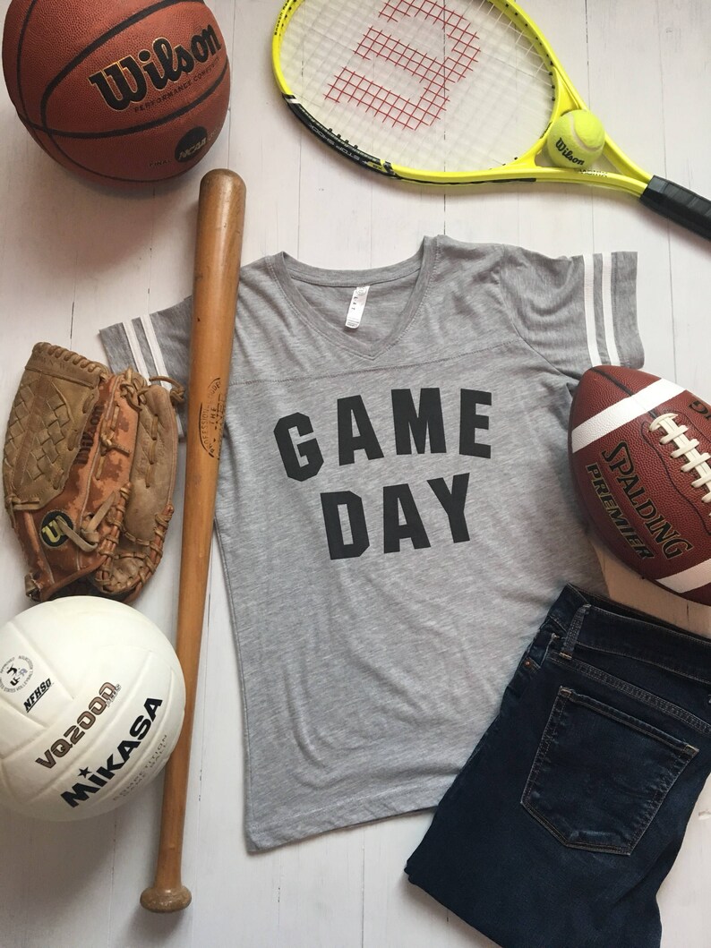 May include: A gray t-shirt with the text "GAME DAY" printed on the front. The shirt has white stripes on the sleeves. The image also includes a baseball bat, a baseball glove, a basketball, a volleyball, a tennis racket, a tennis ball, a football, and a pair of blue jeans.
