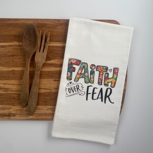 May include: White kitchen towel with the text "Faith over Fear" printed in black. The letters are decorated with a floral pattern in shades of pink, green, yellow, and blue.