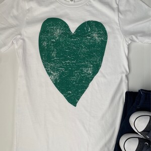 Könnte beinhalten: Weißes T-Shirt mit einem großen, abgenutzten grünen Herz-Motiv.