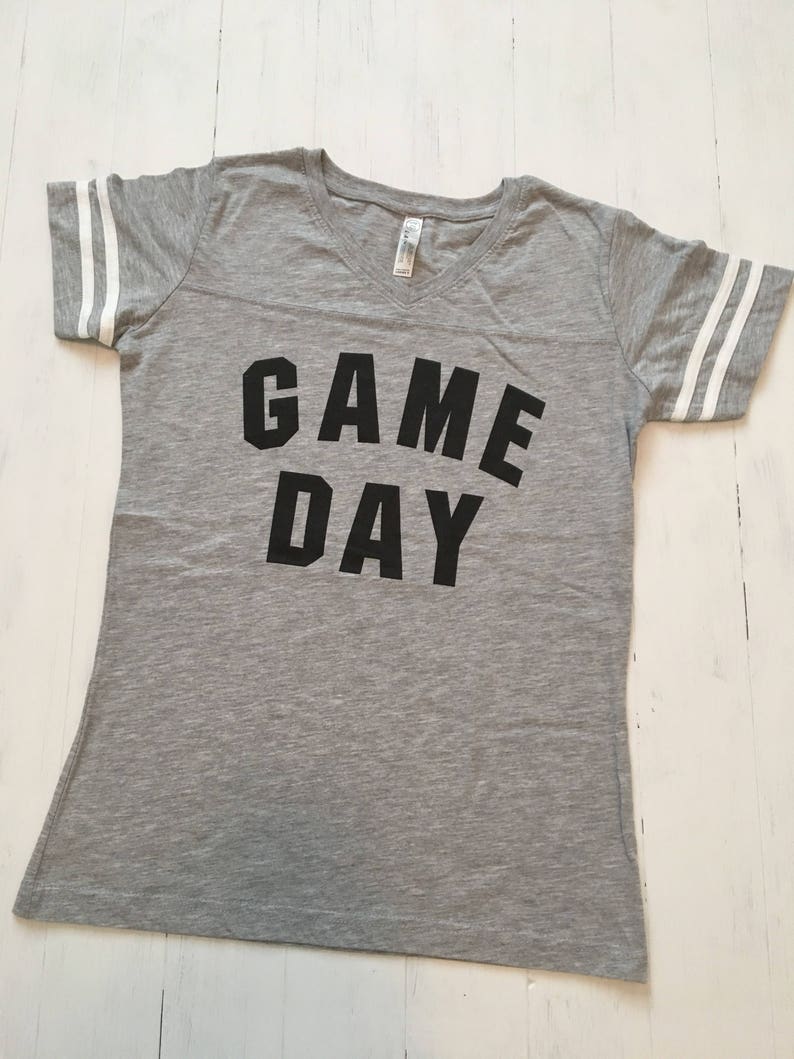 May include: Gray V-neck t-shirt with white stripes on the sleeves and the text "GAME DAY" printed in black.