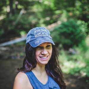 May include: A woman wearing a blue denim baseball cap with the words "Explore" and a mountain range graphic. She is smiling and has long brown hair.