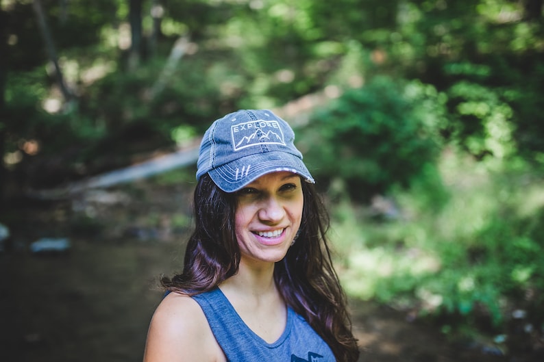 May include: A woman wearing a blue denim baseball cap with the words "Explore" and a mountain range graphic. She is smiling and has long brown hair.