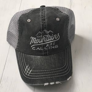 Puede incluir: Una gorra de camionero negra y gris desgastada con un dise&ntilde;o bordado en blanco que dice "The Mountains Are Calling".