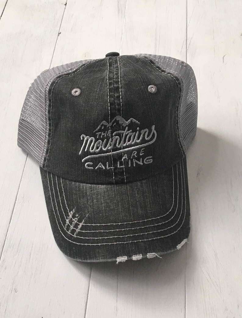 Puede incluir: Una gorra de camionero negra y gris desgastada con un dise&ntilde;o bordado en blanco que dice "The Mountains Are Calling".