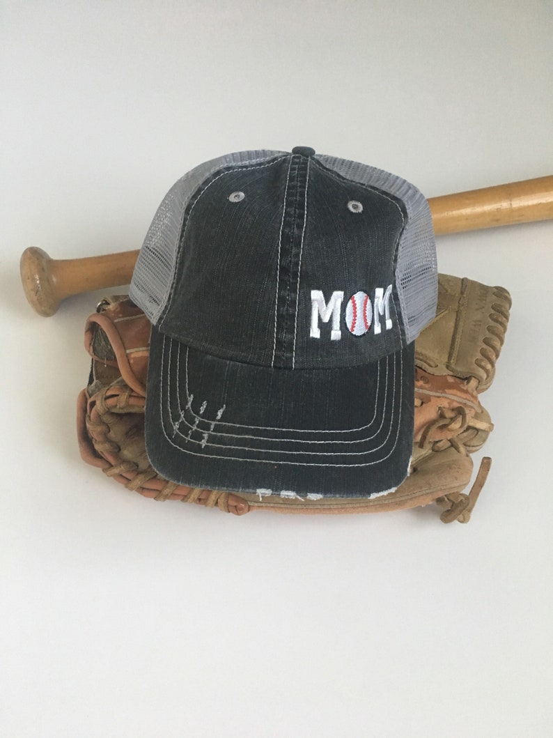May include: A black and grey baseball cap with a white mesh back. The cap has the word "MOM" embroidered on the front with a baseball graphic in the "O".