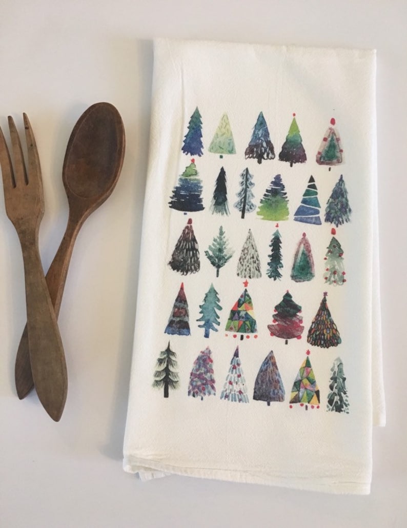 K&ouml;nnte beinhalten: Ein wei&szlig;es Geschirrtuch mit einem farbenfrohen Aquarelldesign aus verschiedenen Weihnachtsb&auml;umen. Die B&auml;ume sind in Blaut&ouml;nen, Gr&uuml;n, Rot und Gelb gemalt.