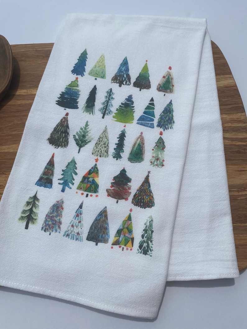 K&ouml;nnte beinhalten: Ein wei&szlig;es K&uuml;chentuch mit einem bunten Muster aus verschiedenen Weihnachtsb&auml;umen. Die B&auml;ume sind in Gr&uuml;n-, Blau-, Rot- und Gelbt&ouml;nen bemalt.