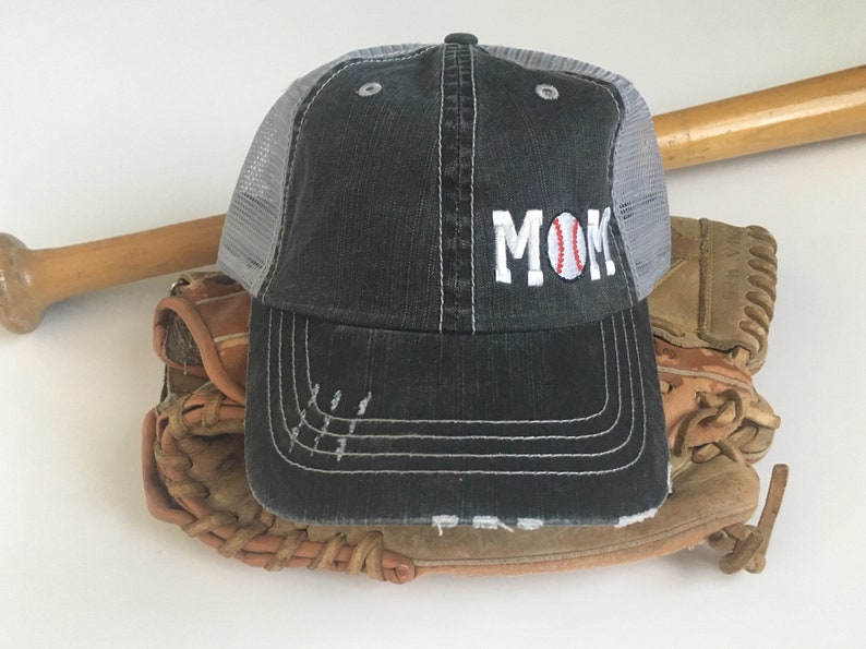 May include: A black and grey baseball cap with a white mesh back. The cap has the word "MOM" embroidered on the front with a baseball in the "O".