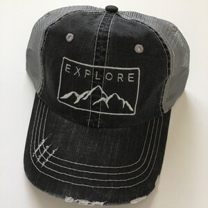 May include: A black and gray distressed trucker hat with a white embroidered mountain range and the word "EXPLORE" in a rectangle.
