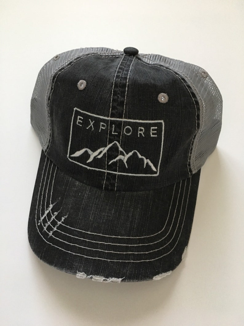 May include: A black and gray distressed trucker hat with a white embroidered mountain range and the word "EXPLORE" in a rectangle.