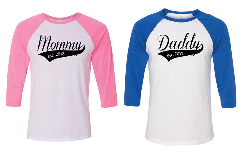 Puede incluir: Dos camisas de estilo b&eacute;isbol con mangas de colores contrastantes. La camisa de la izquierda tiene mangas rosas y dice "Mommy Est. 2018" en texto negro. La camisa de la derecha tiene mangas azules y dice "Daddy Est. 2018" en texto negro.