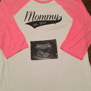 Puede incluir: Una camiseta de b&eacute;isbol blanca y rosa con el texto "Mommy Est. 2017" impreso en la parte delantera. Una imagen de ultrasonido en blanco y negro se coloca sobre la camiseta.