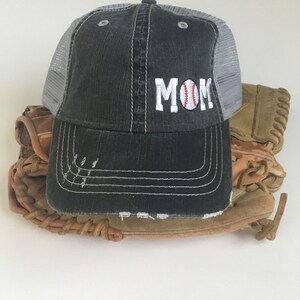 May include: A black and grey baseball cap with a white mesh back. The cap has the word "MOM" embroidered on the front with a red and white baseball graphic.